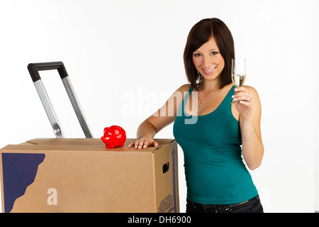 Giovane donna con una scatola di movimentazione, un salvadanaio e un bicchiere di champagne in mano Foto Stock