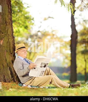 Sorridente senior gentleman seduto su un'erba la lettura di un quotidiano in un parco in autunno Foto Stock