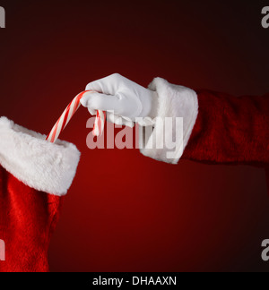 Primo piano di Santa Claus collocando un candy cane in una vacanza calza. Formato quadrato su una luce di colore rosso scuro dello sfondo spot. Foto Stock