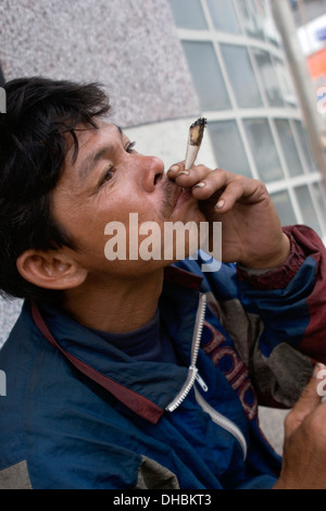 Un povero uomo senza tetto è fumare una casa sigaretta laminati su una strada di città in Khorat, Thailandia. Foto Stock