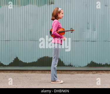 Una ragazza di 10 anni in possesso di un violino sotto il suo braccio, in piedi su una strada. Foto Stock