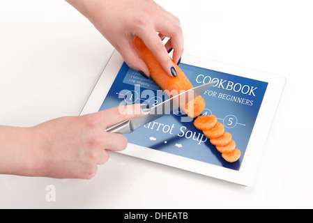 Le mani delle donne è tagliare la carota in un computer tablet, su cui vi è una ricetta di zuppa di carota. Foto Stock