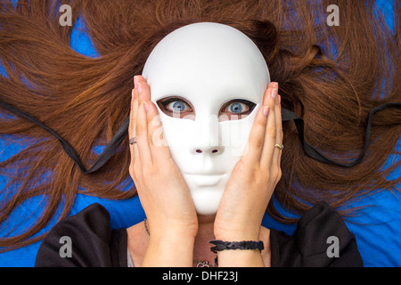 Ragazza brunetta con occhi blu con occhi spaventati che indossa una maschera bianca con un grin creepy fotografato su uno sfondo blu mani sul viso Foto Stock