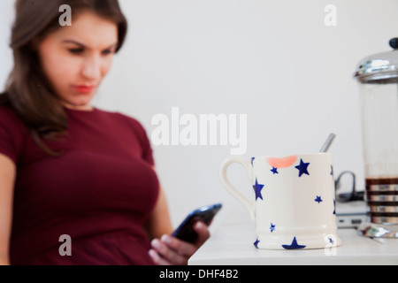 Giovane donna tramite telefono cellulare con tazza da caffè in primo piano Foto Stock