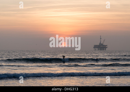 Vista di un surfista californiano stagliano contro il sole al tramonto, con una piattaforma off-shore visibile in distanza. Foto Stock