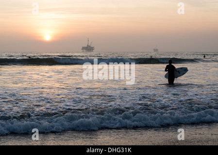 Vista di un surfista californiano stagliano contro il sole al tramonto, con una piattaforma off-shore visibile in distanza. Foto Stock