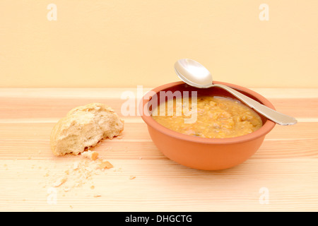 Cucchiaio appoggiato su di una ciotola di zuppa, con la metà di un cilindro di pane croccante e briciole sul tavolo Foto Stock