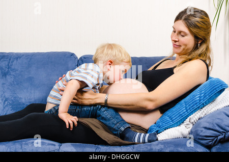 Carino piccolo ragazzo baciare pancia del suo sorridente madre gravida sul divano Foto Stock