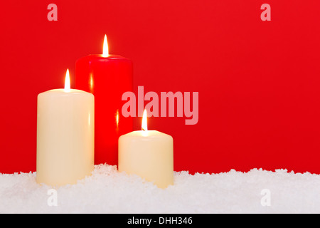 Tre candele di Natale seduti sulla neve contro uno sfondo di colore rosso. Foto Stock