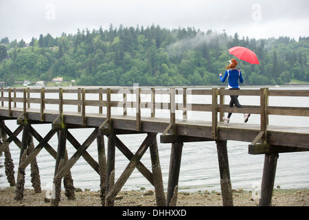 Ragazza adolescente in esecuzione con ombrello sul molo, Bainbridge Island, Washington, Stati Uniti d'America Foto Stock