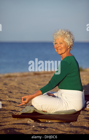 Felice vecchia donna andando esercizio di rilassamento mentre è seduto sulla spiaggia sabbiosa. Senior caucasian praticare yoga dall'oceano in mattinata Foto Stock