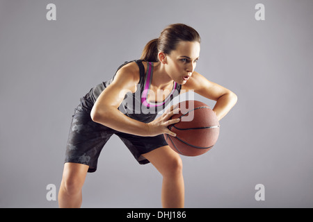 Femmina giovane giocatore di basket in uniforme che passa una palla da basket. Donna in abbigliamento sportivo di giocare a basket e guardando lontano su grigio Foto Stock