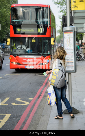 In attesa del bus: giovani femmine passeggero in un London bus stop tiene fuori il suo biglietto o abbonamento al segnale per il bus stop; donna " commuter "; bus rosso Foto Stock