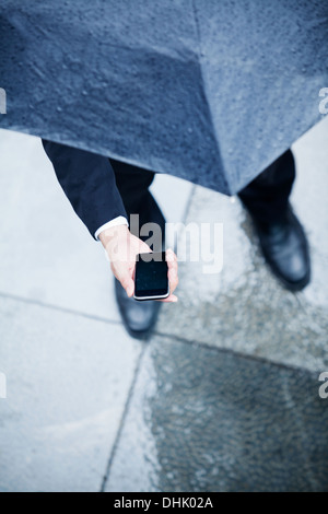 Elevato angolo di visione dell uomo d affari con ombrello e guardando il suo telefono sotto la pioggia Foto Stock