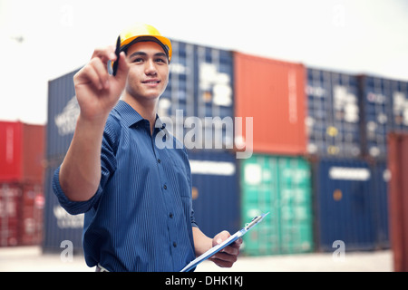 Sorridente giovane ingegnere in protezione usura di lavoro in un cantiere di spedizione esaminando cargo Foto Stock