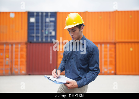 Sorridente giovane ingegnere in protezione usura di lavoro in un cantiere di spedizione esaminando cargo e iscritto negli appunti Foto Stock