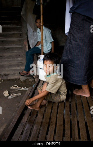 Un giovane bambino operaio ragazzo che vivono in condizioni di povertà è seduta su di un letto di legno in un tugurio sgangherato in Kampong Cham, Cambogia. Foto Stock