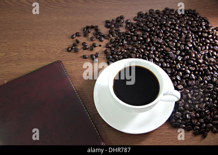 La messa a fuoco delle immagini bianco tazza di caffè e un libro sul tavolo di legno vecchio vicino i chicchi di caffè. Foto Stock