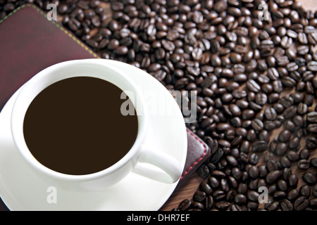 La messa a fuoco delle immagini bianco tazza di caffè e un libro sul tavolo di legno vecchio vicino i chicchi di caffè. Foto Stock