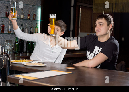 Due giovani uomini seduti al bancone del bar in un club di elevare i loro bicchieri da birra in aria in alto in un toast come essi celebrare insieme. Foto Stock