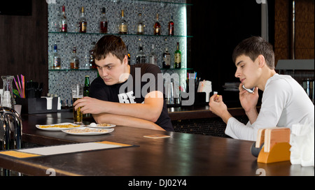 Due uomini amici divertendosi avente un bicchiere di birra al bar con uno occupato avente una conversazione sul suo telefono cellulare Foto Stock