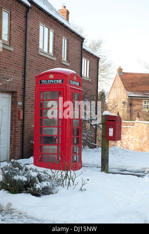 Un red UK Royal Mail post scatola fissata ad un palo di legno nella neve accanto ad un tradizionale rosso BT casella Telefono. Foto Stock