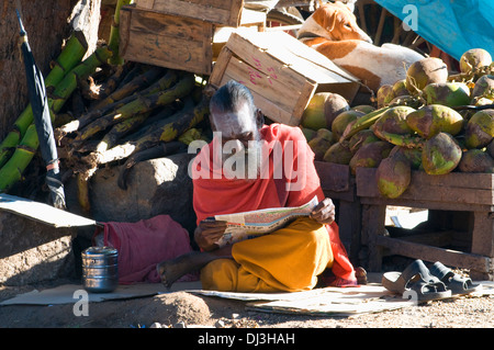 Sadhu, Indiana uomo santo e renunciant siede leggendo il giornale a coconut stand mentre il cane del villaggio si crogiola al sole dietro Foto Stock