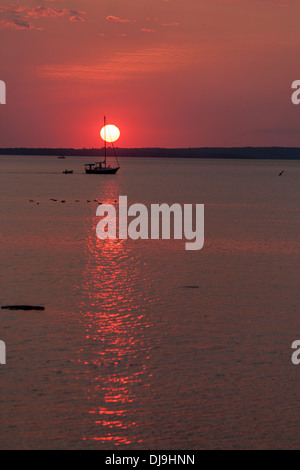 Attraverso il tramonto. Una barca a vela con le sue vele vele arrotolata attraverso la luce di un sole di setting. Foto Stock