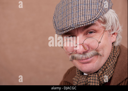 Sorridente dai capelli grigi vecchio uomo che indossa il tappo piatto,bicchieri e comodi sciarpa, contro uno sfondo di colore marrone. Foto Stock