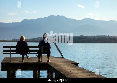 L uomo e la donna seduta su una panca in legno posto sul molo che si affaccia su lago di Alpi Bavaresi, Chiemsee, Chiemgau, Alta Baviera Foto Stock