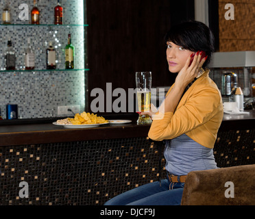 Giovane donna seduta a un banco di bar in una discoteca ascoltare una chiamata sul suo telefono cellulare guardando la telecamera Foto Stock