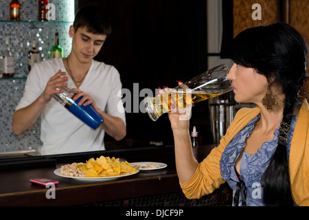 Barman lavorando dietro un banco di bar in un night club cocktail di miscelazione controllata da una giovane donna sorseggiando una pinta di birra Foto Stock