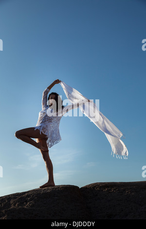 Modello femminile sventolare una lunga sciarpa bianca in aria contro un profondo cielo blu mentre si fermò a piedi nudi su una roccia che indossa bikini bianco Foto Stock