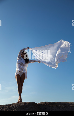 Modello femminile sventolare una lunga sciarpa bianca in aria contro un profondo cielo blu mentre si fermò a piedi nudi su una roccia che indossa bikini bianco Foto Stock