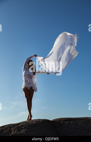 Modello femminile sventolare una lunga sciarpa bianca in aria contro un profondo cielo blu mentre si fermò a piedi nudi su una roccia che indossa bikini bianco Foto Stock