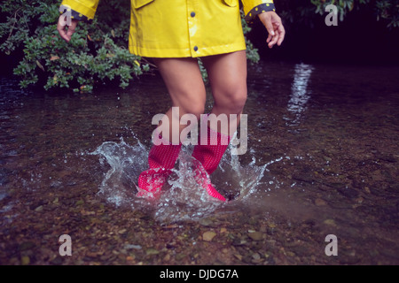 Donna in impermeabile giallo e rosso gumboots saltando in acqua Foto Stock