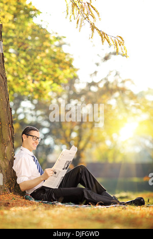 Giovane uomo con gli occhiali seduto su un prato verde e la lettura di un quotidiano in un parco Foto Stock