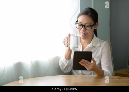 Una bella donna asiatica detiene un ipad, alla ricerca e alla lettura di bere il caffè nella sua casa. Foto Stock