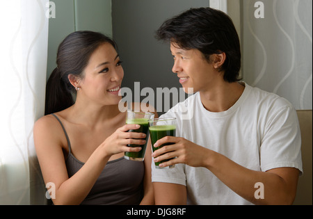 Una giovane coppia asiatica sollevando un bicchiere di succo verde e sorridente ad ogni altro, celebrando il benessere e buona salute Foto Stock