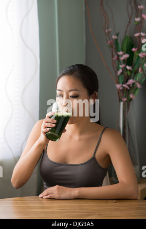 Una giovane donna asiatica di bere un bicchiere di verde succo di verdura, wheatgrass, seduti a un tavolo nella sua casa. Foto Stock