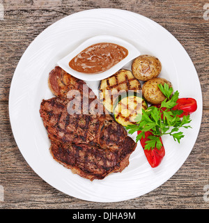 Bistecche alla griglia, patate al forno e verdure sulla piastra bianca su sfondo di legno. Foto Stock