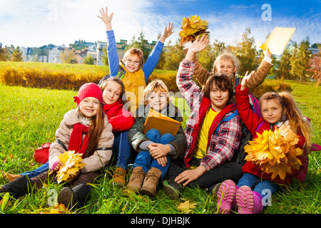 Un folto gruppo di bambini, amici, i ragazzi e le ragazze circa dieci anni, seduta in erba in autunno vestiti con le foglie di acero mazzi e carte dopo la scuola con le mani sollevate Foto Stock