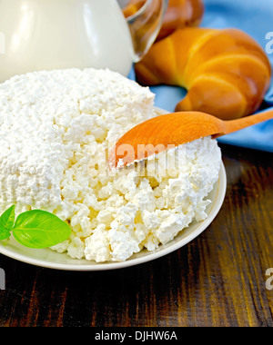 Il formaggio in un piattino con basilico, il cucchiaio di legno, due ciambelle di pane, una brocca di latte, il tovagliolo sulla tavola di legno Foto Stock