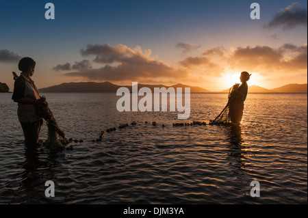 Sunset silhouettes due donne che assumono nella loro rete da pesca fuori del villaggio di Tovu in Totoya caldera del. Lau Isole Figi. Foto Stock