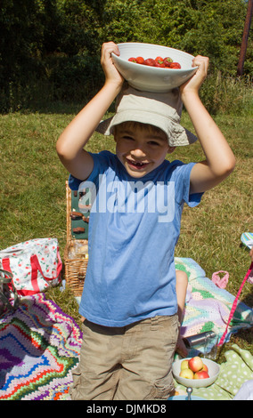 Un giovane ragazzo che indossa una maglietta blu e cappello per il sole tenendo una tazza di fragole e sorridente alla fotocamera, su un picnic in Somerset Foto Stock