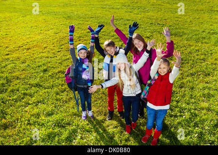 Gruppo di felice teen età i bambini della scuola in piedi sul prato con sorriso sui volti mani di sollevamento, vista dall'alto, indossando vestiti caldi Foto Stock