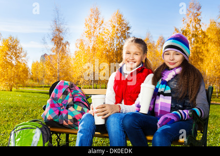 Due ragazze felici 11 anni seduto sul banco in autunno park con zaini e caffè Foto Stock