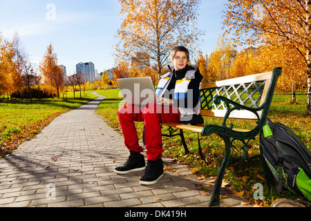 Dieci anni vecchio ragazzo seduto sul banco in autunno park lavorando sul computer portatile esterno sulla giornata di sole Foto Stock