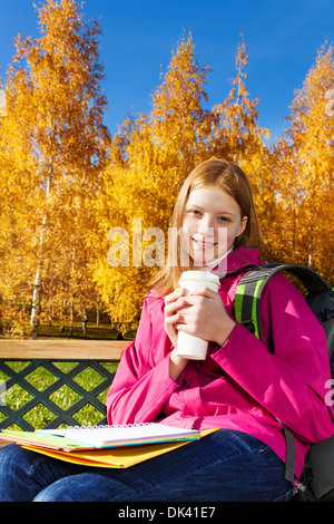 Bella teen 14 anni di scuola vecchia ragazza seduta sul banco in autunno park azienda caffè e libri di testo Foto Stock