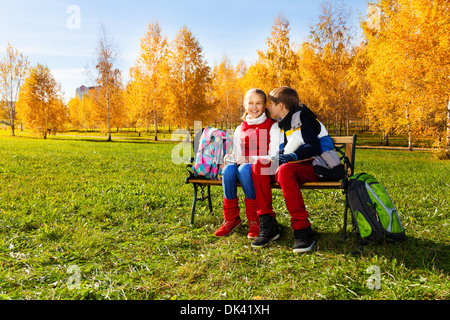 Accoppiare i bambini, ragazzo sussurra alla ragazza seduta sul banco in autunno park con zaini che stabilisce nelle vicinanze Foto Stock